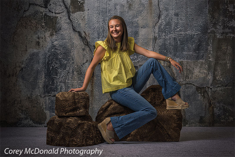Textured Silver Industrial Wall photography backdrop shown with a senior girl in a yellow ruffle-sleeve top and blue flared jeans seated on the brown rocks with one arm resting on her knee on faux rock props CP-8807