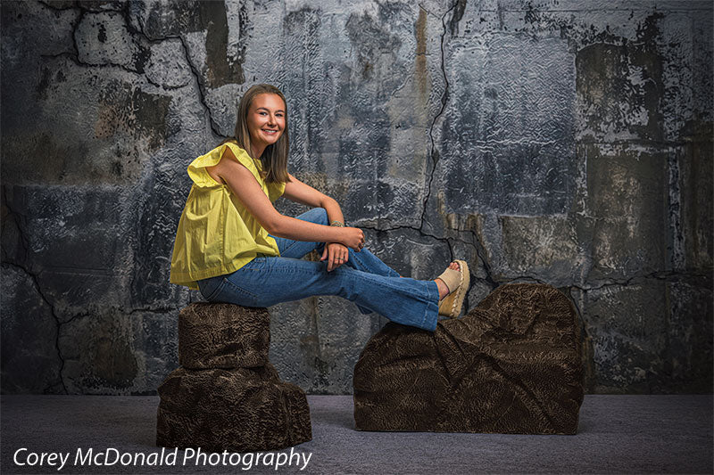 Textured Silver Industrial Wall photography backdrop shown with a senior girl in a yellow ruffle-sleeve top and blue flared jeans seated on the brown rocks in profile with a hand on her knee on faux rock props CP-8807