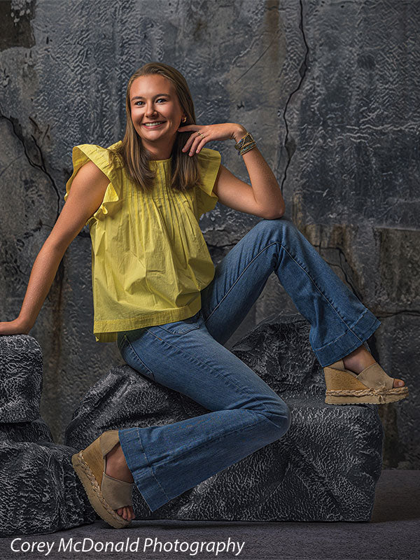 Textured Silver Industrial Wall photography backdrop shown with a senior girl in a yellow ruffle-sleeve top and blue flared jeans seated on the rocks with a hand near her chin on faux rock props CP-8807