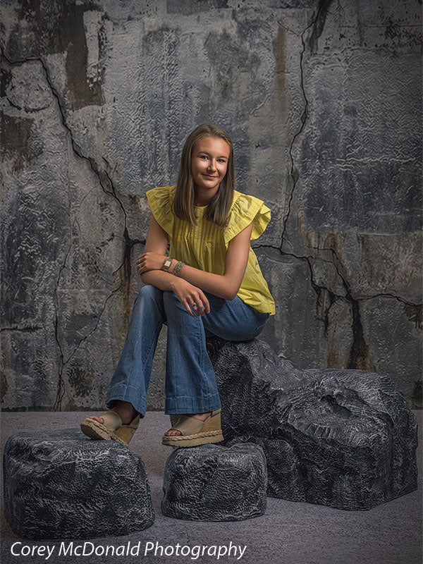 Textured Silver Industrial Wall photography backdrop shown with a senior girl in a yellow ruffle-sleeve top and blue flared jeans seated on the rocks with hands clasped over one knee on faux rock props CP-8807