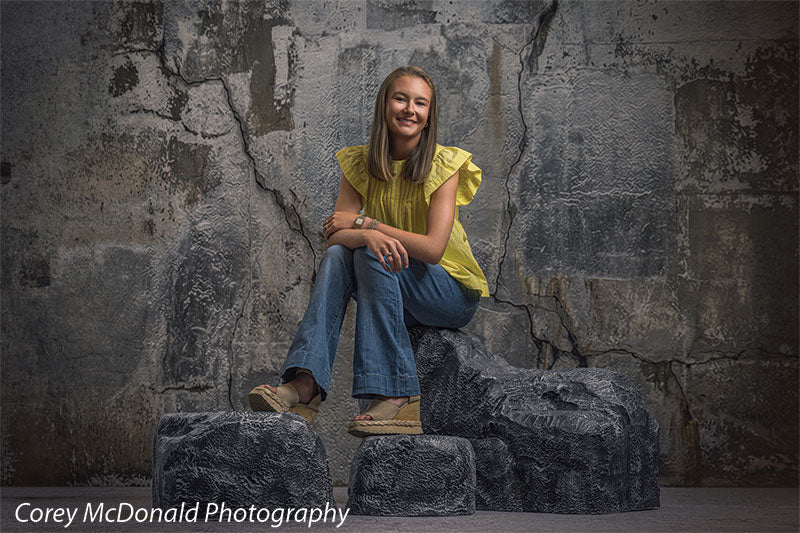 Textured Silver Industrial Wall photography backdrop shown with a senior girl in a yellow ruffle-sleeve top and blue flared jeans seated on the rocks with one foot resting forward on faux rock props CP-8807