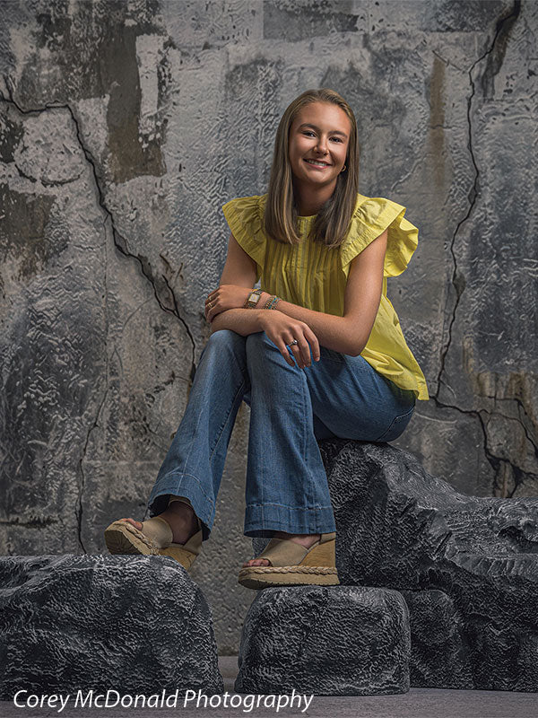 Textured Silver Industrial Wall photography backdrop shown with a senior girl in a yellow ruffle-sleeve top and blue flared jeans seated on the stacked rocks with one ankle crossed over her knee on faux rock props CP-8807