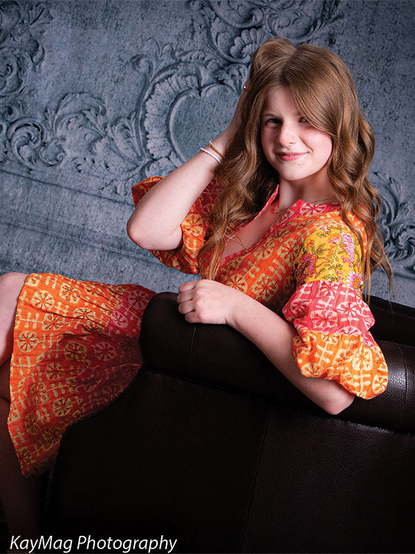 Girl with long wavy hair posing on a leather chair against a baroque-inspired gray-blue wall backdrop, great for timeless senior or youth portraits.
