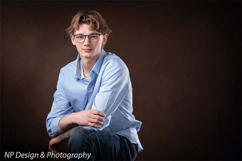 Toasted chestnut old masters photography backdrop shown with a senior boy in a light blue shirt seated with his arms crossed on his knee OM-2622