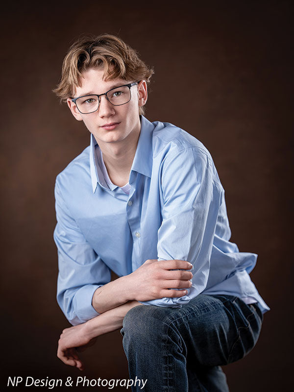 Toasted chestnut old masters photography backdrop shown with a senior boy in a light blue shirt seated with his hands clasped on his knee OM-2622