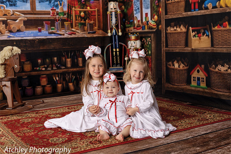 Toy Maker Santa's workshop craft-table photography backdrop shown as part of a Santa's workshop Christmas corner set with three young girls in white Christmas nightgowns seated on the workshop rug, wide view CP-8943