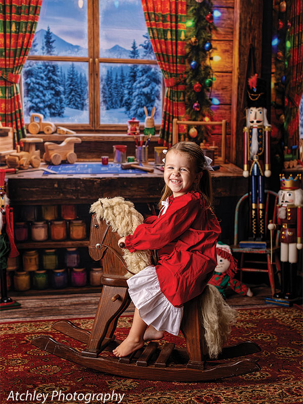 Toy Maker Santa's workshop craft-table photography backdrop shown as part of a Santa's workshop Christmas corner set with a young girl in a red dress on a wooden rocking horse CP-8943