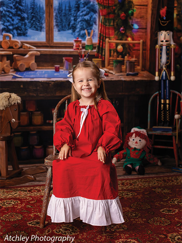 Toy Maker Santa's workshop craft-table photography backdrop shown as part of a Santa's workshop Christmas corner set with a young girl in a red Christmas gown seated on an antique chair CP-8943