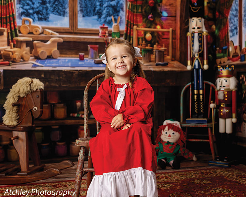 Toy Maker Santa's workshop craft-table photography backdrop shown as part of a Santa's workshop Christmas corner set with a young girl in a red Christmas gown seated on an antique chair, wide view CP-8943