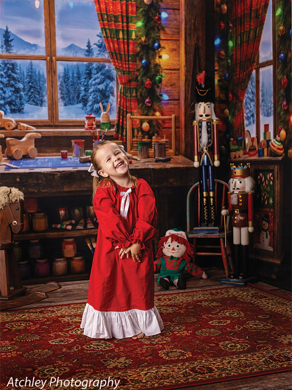 Toy Maker Santa's workshop craft-table photography backdrop shown as part of a Santa's workshop Christmas corner set with a young girl in a red Christmas gown standing and laughing CP-8943