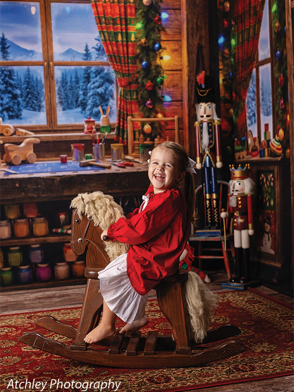 Toy Maker Santa's workshop craft-table photography backdrop shown as part of a Santa's workshop Christmas corner set with a young girl in a red dress laughing on a wooden rocking horse CP-8943