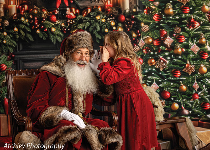 Young girl in a red dress whispering into Santa Claus’s ear while he sits in an armchair, set against a traditional Christmas tree and fireplace backdrop decorated with garland, wreath, candles, and wrapped presents.