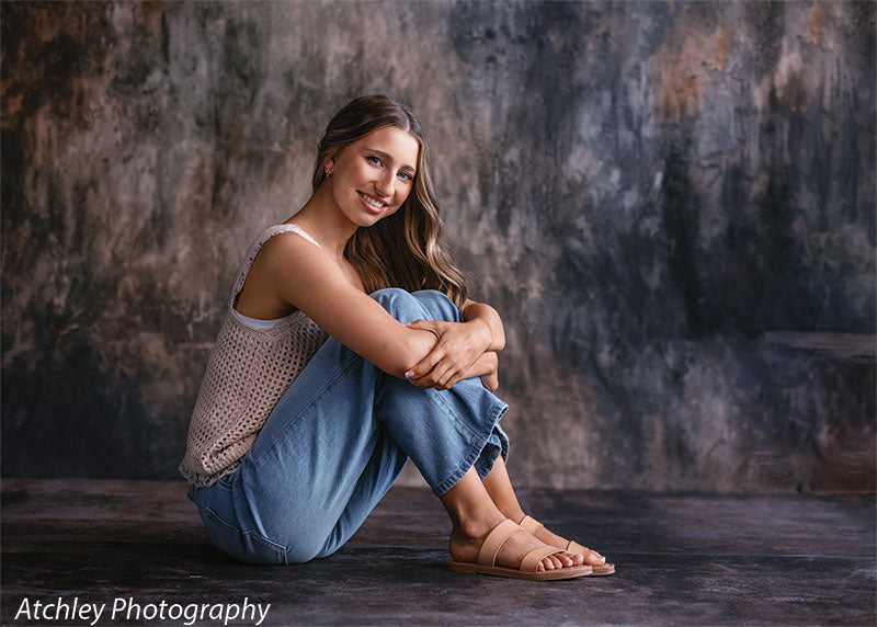 Young woman with long brown hair sitting on a textured concrete floor with her knees pulled in, wearing a cream knit top, light blue jeans, and tan sandals, photographed against a dramatic mottled gray and brown studio wall with the turville concrete floor floordrop extending beneath her.