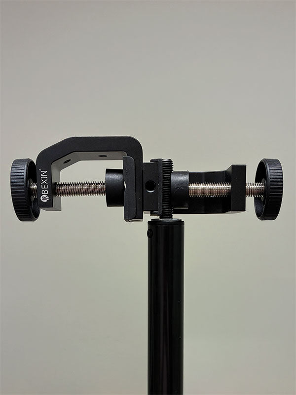 Black metal Twist Flex Clamp shown from the front mounted on top of a light stand pole, with its C-clamp jaw and dual knurled tightening knobs on a threaded crossbar