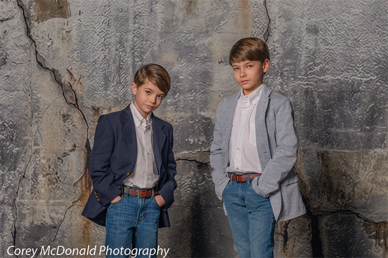 Two boys dressed in white shirts with blue and gray blazers and jeans pose together with hands in pockets in front of a weathered gray concrete wall backdrop featuring visible cracks and industrial texture.