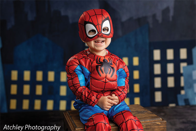 A toddler wearing a red and blue superhero costume and mask sits on a wooden box with hands together, smiling in front of an illustrated city night backdrop with lit windows.
