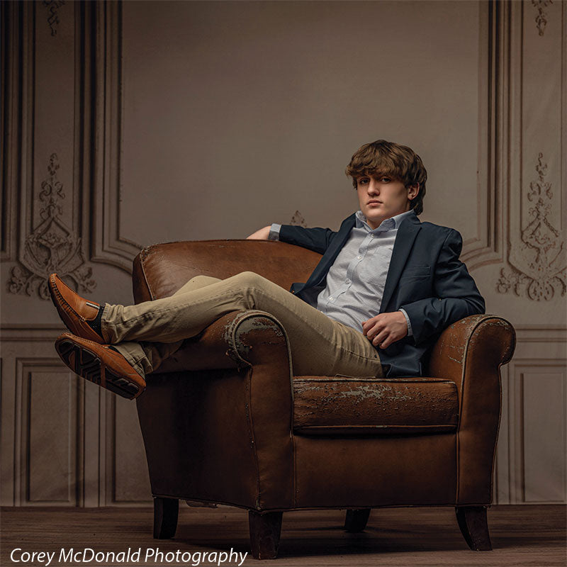 Teen boy with wavy light brown hair wearing a blue blazer and khaki pants reclines casually across a distressed brown leather armchair with one leg stretched out, posed in front of a vintage beige wall backdrop with decorative paneling.