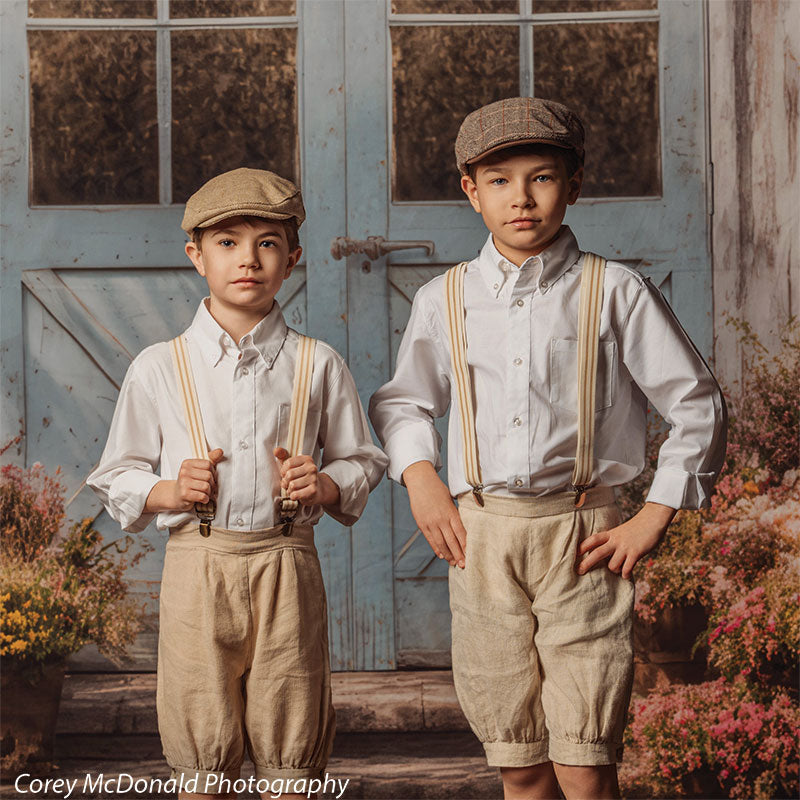 Two young boys wearing white shirts, beige shorts, suspenders, and caps pose in front of a vintage blue wooden door backdrop with rustic paneling and surrounding floral borders, both looking forward with calm expressions.