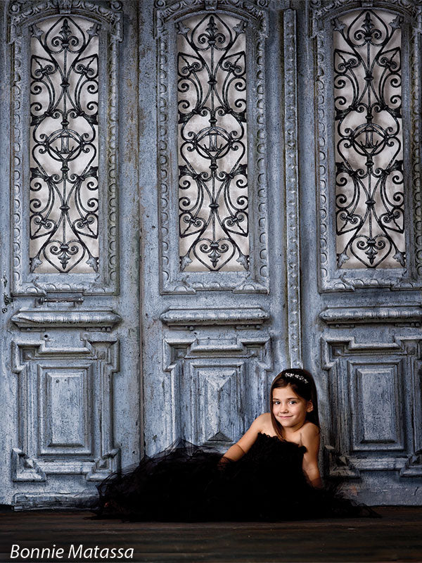 Girl in a black formal gown sitting gracefully in front of an ornate vintage blue door backdrop with iron scrollwork, ideal for elegant children's portraits.
