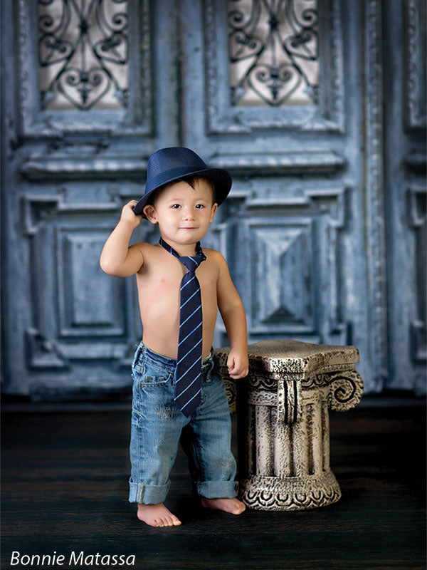 Toddler boy wearing a fedora and tie poses confidently in front of a blue antique door backdrop with iron panels, great for stylized toddler portraits and vintage themes.