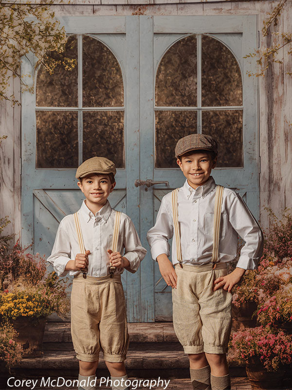 Two young boys dressed in white shirts, beige shorts, suspenders, and caps stand side by side smiling in front of a vintage blue wooden door backdrop with arched window panels and surrounding floral arrangements.