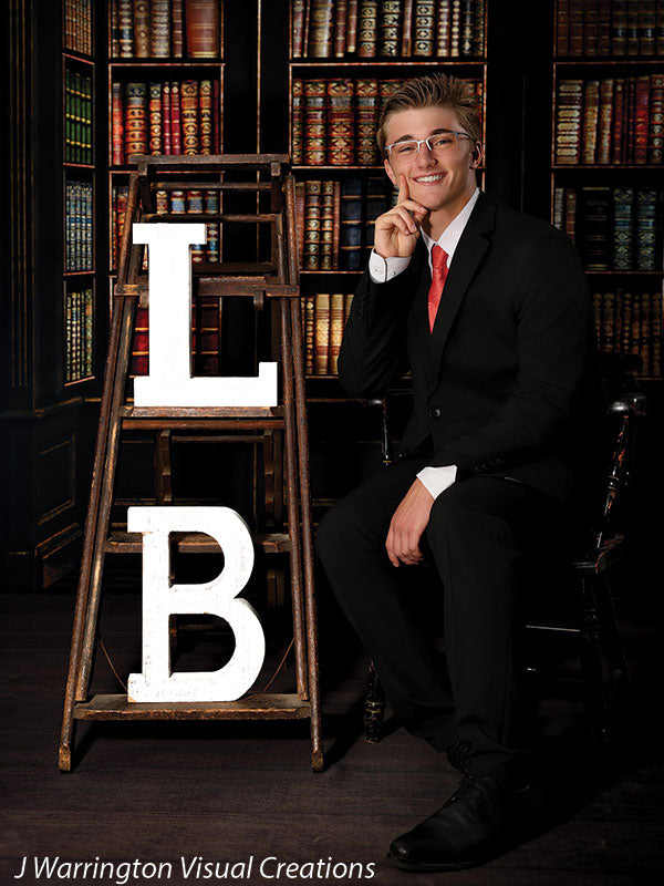 Senior boy smiling beside wooden step ladder with initials “L” and “B” in front of a vintage bookcase backdrop, styled for graduation or senior portrait sessions with props.