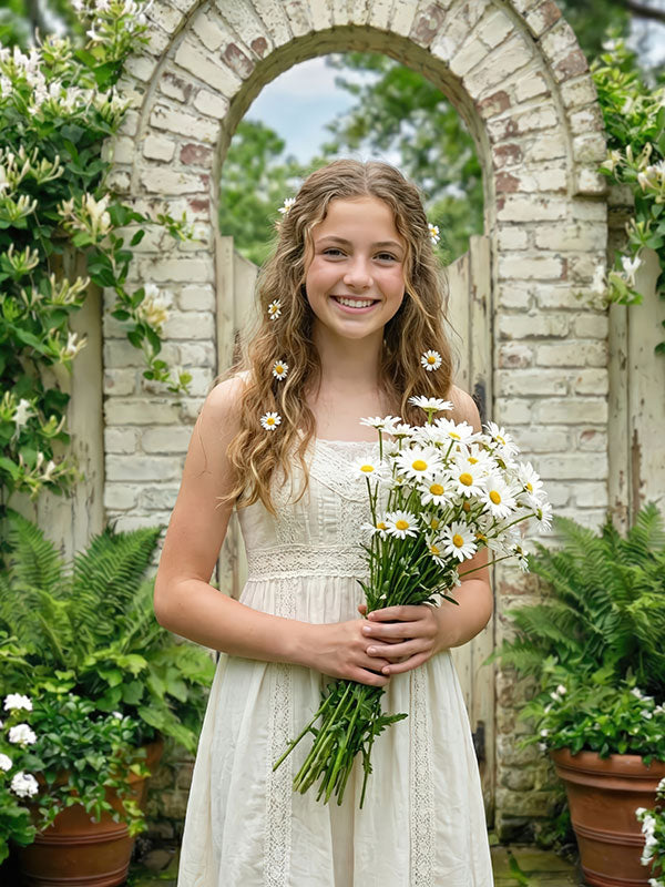 Young girl wearing a white lace dress holds a bouquet of daisies while smiling in front of a vintage brick garden arch backdrop surrounded by green plants and potted foliage.