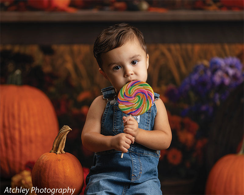 Toddler in denim overalls holding a rainbow swirl lollipop while sitting near pumpkins and autumn florals, set against a vintage carnival fairground backdrop with a lit carousel, baskets of gourds, and warm fall foliage.