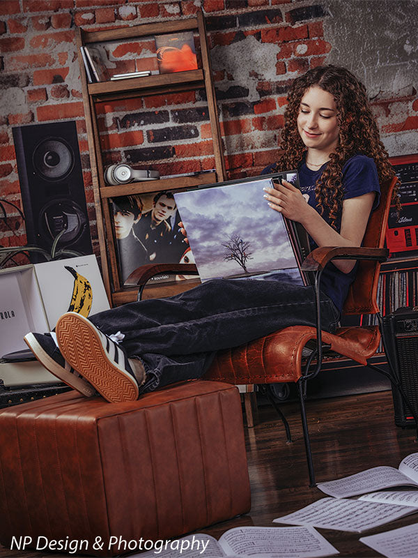 Vintage exposed brick music studio photography backdrop shown with a senior girl reclining in a leather chair with an album print in a vertical portrait CP-8630