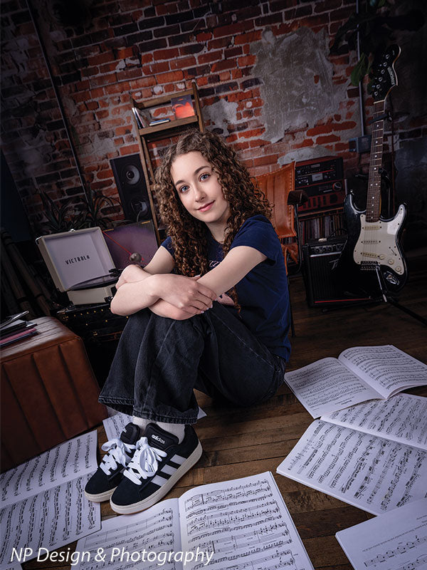 Vintage exposed brick music studio photography backdrop shown with a senior girl seated on the floor hugging her knees among scattered sheet music CP-8630