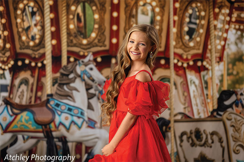 Young girl with long wavy blonde hair wearing a red high-low dress, posing gracefully in front of a vintage fairground carousel portrait backdrop with painted horses, mirrors, and illuminated bulbs.