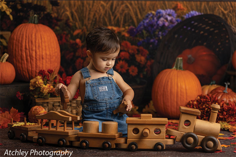 Toddler sitting on a small stool holding a colorful swirl lollipop, surrounded by pumpkins and scattered fall leaves, posed in front of a vintage fall fair backdrop with baskets of gourds, flowers, and a lit carousel.
