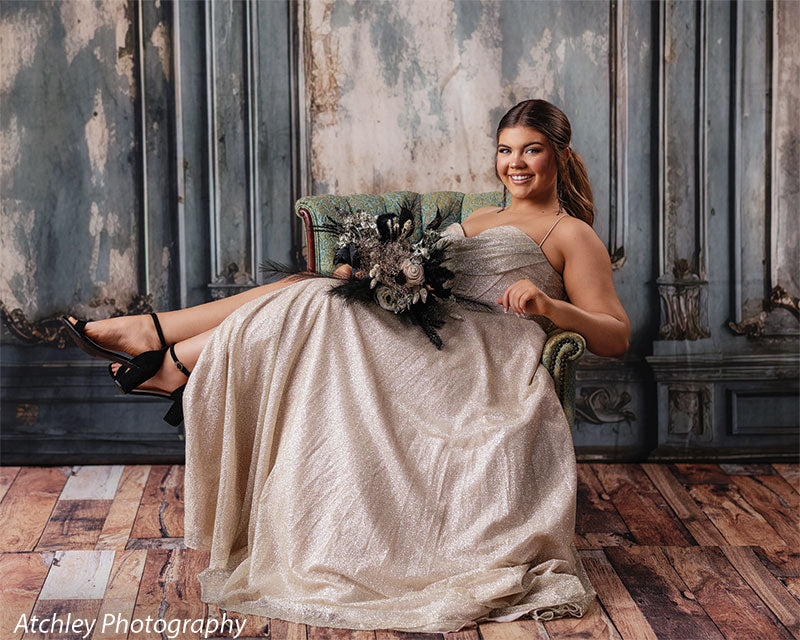 French panel-inspired studio backdrop with blue distressed panels and a wood floor, female senior reclining in a gold gown on an antique chair, perfect for sophisticated photography.