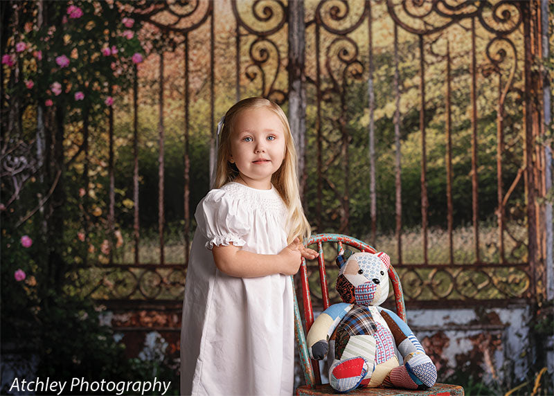 A young girl wearing a white dress stands beside a small chair holding a patchwork teddy bear, posed in front of a vintage iron garden gate backdrop with soft greenery.
