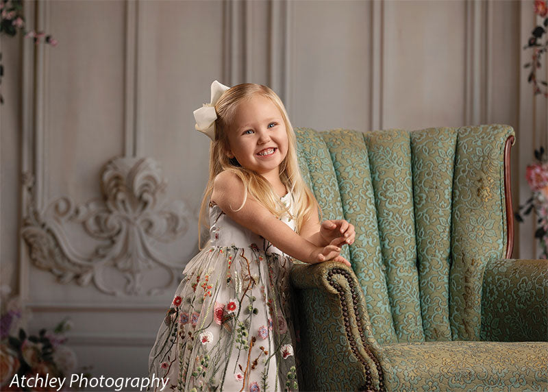 A young girl in a floral embroidered dress leans on a green upholstered chair, smiling, in front of a vintage ornate wall backdrop framed with flowers.
