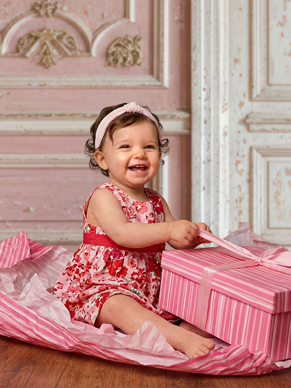 Baby wearing a pink floral dress and headband sits on the floor smiling while holding a pink gift box, posed in front of a pastel pink vintage painted door backdrop with decorative paneling.