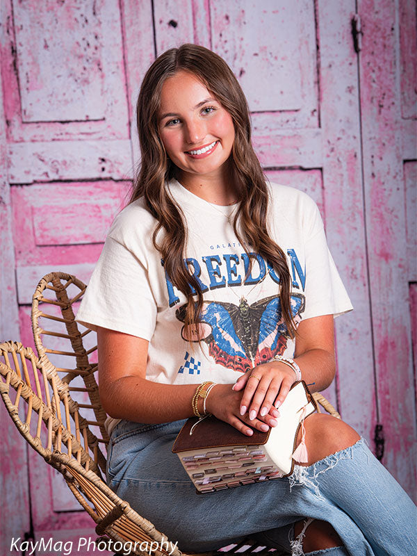 Teen girl holding a book while seated on a wicker chair in front of a vintage pink paneled backdrop, suited for styled studio portrait scenes.