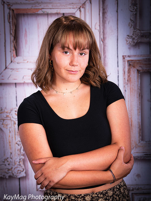 Confident teen girl posing with arms crossed in front of a rustic whitewashed wooden backdrop with vintage picture frames, ideal for studio senior portraits.