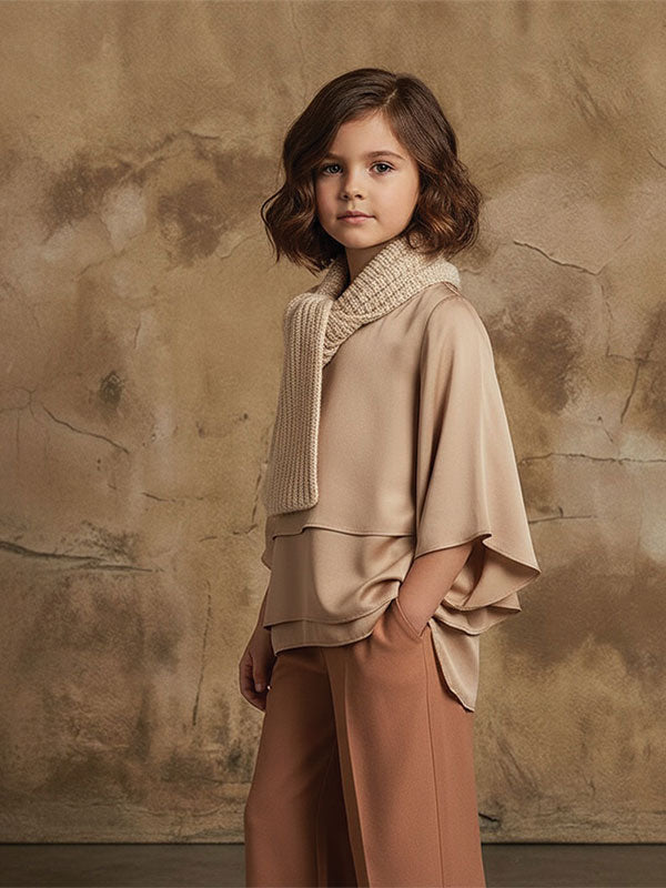 Young girl with short brown hair wearing a beige top, matching scarf, and brown pants stands with one hand in her pocket in front of a warm beige textured plaster wall backdrop.