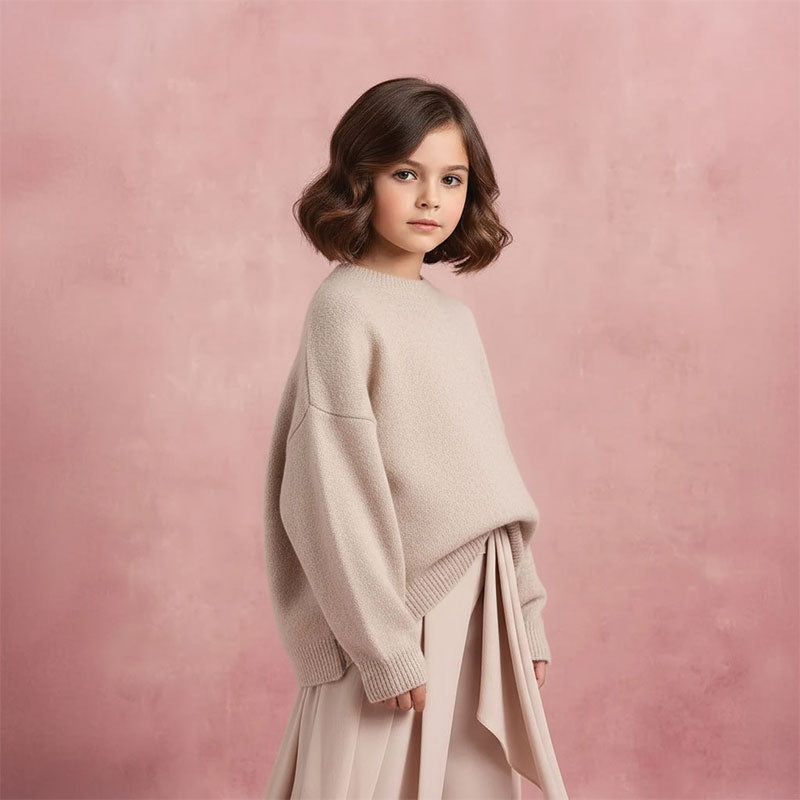 Young girl with brown bob hairstyle wearing a neutral beige sweater and skirt stands facing forward with a calm expression against a warm blush textured portrait backdrop.
