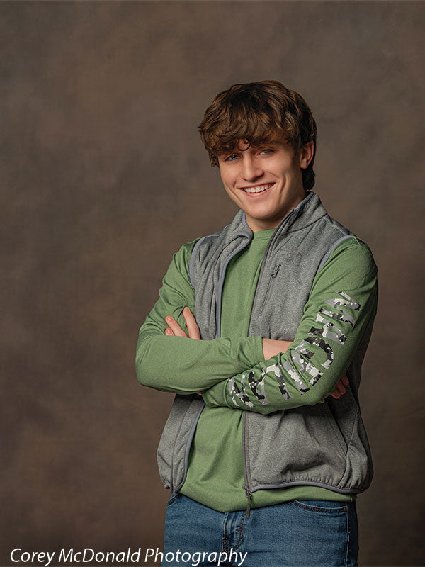 Teen boy with wavy brown hair wearing a green long sleeve shirt, gray vest, and blue jeans stands with arms crossed and smiling in front of a warm brown mottled studio backdrop with soft blended texture.