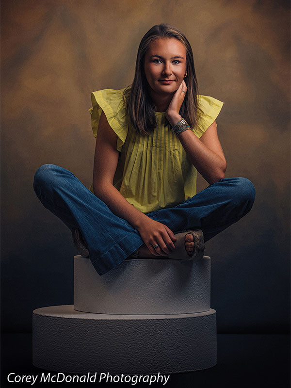 Warm Elegance warm brown mottled texture photography backdrop shown with a senior girl in a yellow ruffle-sleeve top and blue flared jeans seated cross-legged on the pedestals with her chin resting on her hand on circular foam posing pedestals OM-2459