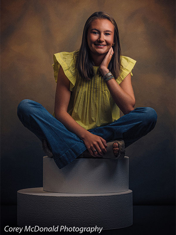 Warm Elegance warm brown mottled texture photography backdrop shown with a senior girl in a yellow ruffle-sleeve top and blue flared jeans seated cross-legged on the stacked pedestals with a bright smile on circular foam posing pedestals OM-2459