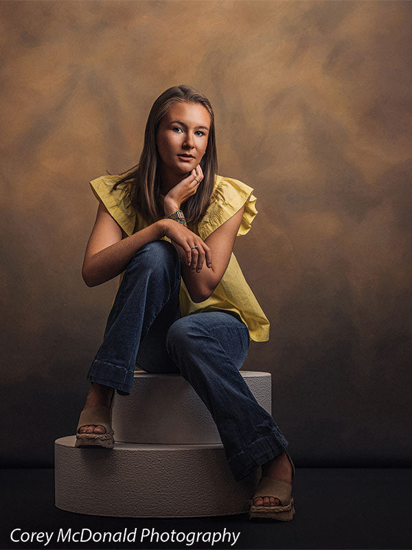 Warm Elegance warm brown mottled texture photography backdrop shown with a senior girl in a yellow ruffle-sleeve top and blue flared jeans seated on the stacked pedestals with a hand on her knee on circular foam posing pedestals OM-2459