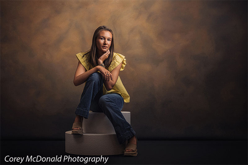Warm Elegance warm brown mottled texture photography backdrop shown with a senior girl in a yellow ruffle-sleeve top and blue flared jeans seated on a pedestal with hands clasped in a wide studio view on circular foam posing pedestals OM-2459