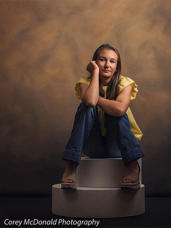 Warm Elegance warm brown mottled texture photography backdrop shown with a senior girl in a yellow ruffle-sleeve top and blue flared jeans seated on a pedestal with her knees drawn up on circular foam posing pedestals OM-2459
