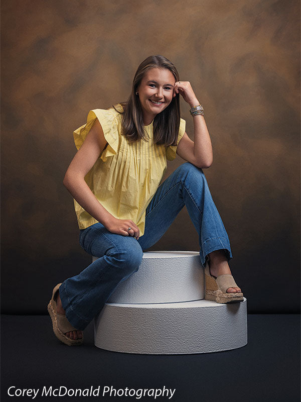 Warm Elegance warm brown mottled texture photography backdrop shown with a senior girl in a yellow ruffle-sleeve top and blue flared jeans seated on a pedestal, smiling with a hand near her face on circular foam posing pedestals OM-2459