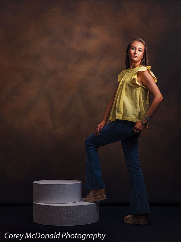 Warm Elegance warm brown mottled texture photography backdrop shown with a senior girl in a yellow ruffle-sleeve top and blue flared jeans standing with one foot on the pedestal in a full-length pose on circular foam posing pedestals OM-2459