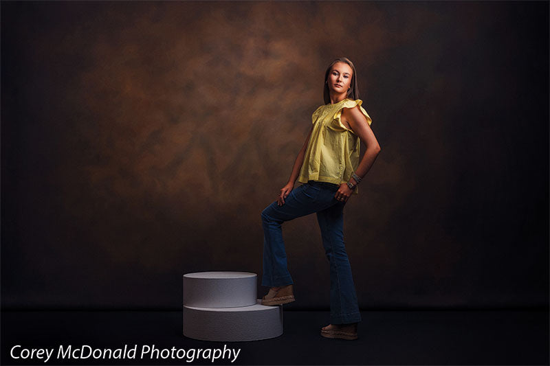 Warm Elegance warm brown mottled texture photography backdrop shown with a senior girl in a yellow ruffle-sleeve top and blue flared jeans standing with one foot on the pedestal and a hand on her hip on circular foam posing pedestals OM-2459