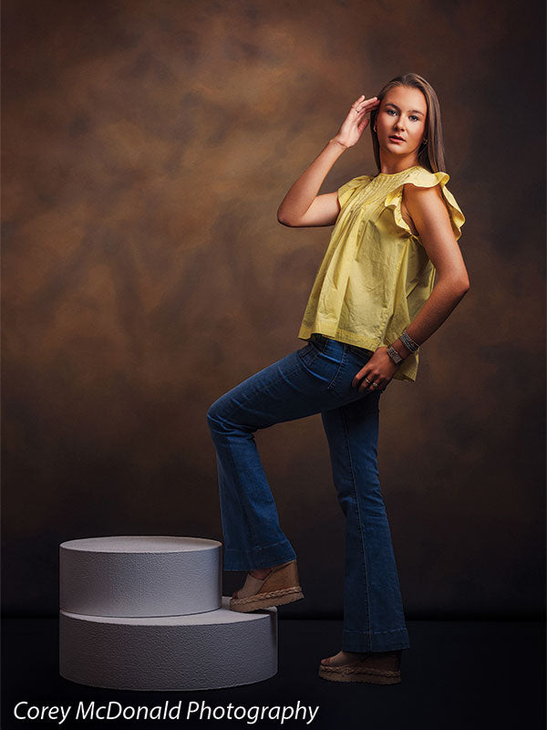 Warm Elegance warm brown mottled texture photography backdrop shown with a senior girl in a yellow ruffle-sleeve top and blue flared jeans standing with a hand in her hair and one foot on the pedestal on circular foam posing pedestals OM-2459